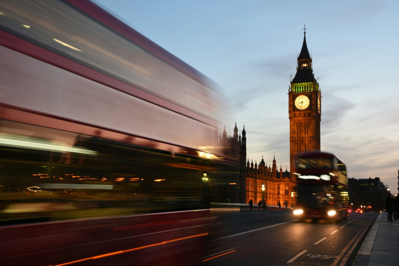 Big Ben illuminated at dusk with blurred red double-decker buses passing by on Westminster Bridge.