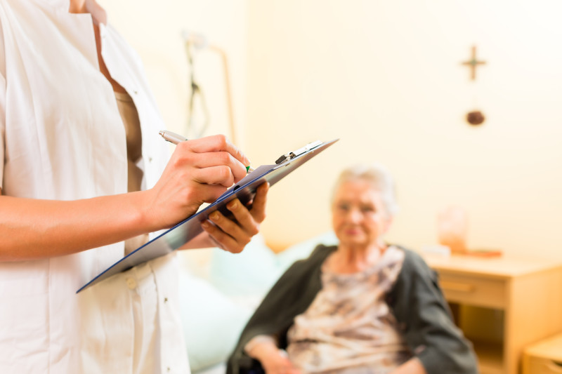 A care worker fills out a clipboard chart in front of an elderly woman seated in a care home room.
