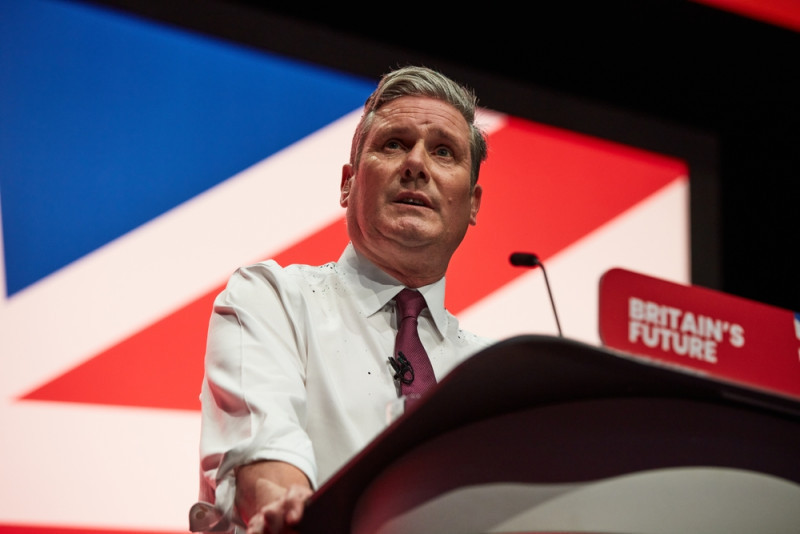 A man with silver hair stands at a podium, speaking into a microphone. He is wearing a white shirt and a maroon tie, with a backdrop featuring the Union Jack and the words "Britain's Future."