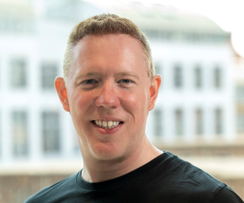 A smiling man with short, light brown hair wearing a black t-shirt stands in front of a blurred urban background.