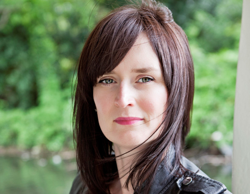 A woman with shoulder-length brown hair and green eyes stands outdoors, looking confidently at the camera. She wears a black leather jacket, and the background features blurred greenery.