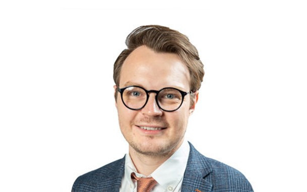 A man with glasses and light brown hair smiles warmly. He is wearing a checked blazer and a tie, posing against a plain white background.
